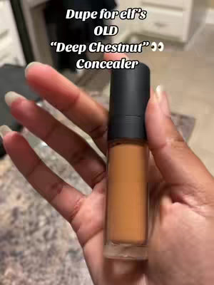 A hand holding a concealer tube against a blurred background. The concealer is a deep brown shade. Text overlayed on the image reads "Dupe for elf's OLD 'Deep Chestnut' Concealer".