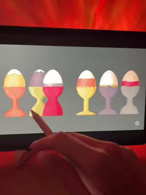 A hand with pink nail polish holds a stylus over an iPad screen. On the screen are six stylized eggs in egg cups of different colors (yellow, purple, red, etc.), each with a white boiled egg on top. The background is a fiery red.