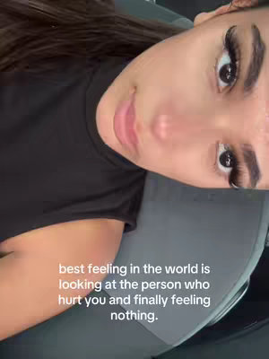A selfie-style shot with a woman looking up towards the camera, framed from a close-up angle. She is wearing a black top, and her face takes up most of the frame revealing her face features like eyelashes and lips. The background is blurred and out of focus. Text is overlaid at the bottom, which is easy to read.