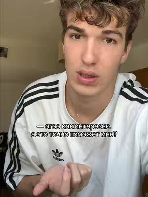 A young man with light brown hair stands in front of a light beige background. He wears a white Adidas t-shirt with black stripes on the sleeves and the Adidas logo in the center. The man is looking at the camera and talking. Overlay text is in Russian.