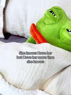 A plush green frog with an orange neck is seen lying in a bed, the frog is tucked under a white blanket, the top of the frog’s head with its black eyes are visible. The text overlayed on the image says, “She knows I love her but I love her more than she knows”.