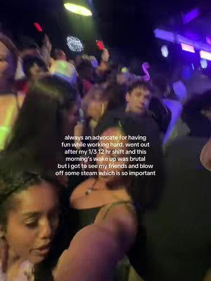 An image of a crowded bar. There is a woman to the left with curly black hair and a black strapless top looking at the camera. The background is dark and blurry, with many people visible and some lights.