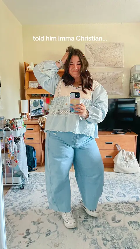A mirror selfie showing a young woman wearing a light blue blouse, light blue jeans, and white sneakers. She is smiling, holding her hair, and holding a phone to take the photo. The background shows a room with a wooden cabinet, a rug, and world map decorations.
