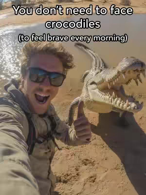 A selfie-style shot of a man, wearing sunglasses and a khaki jacket, smiling and giving a thumbs-up. Beside him is a large crocodile with its mouth wide open. The background shows a beach and water.