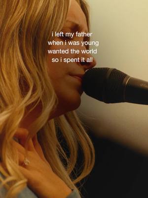A close-up shot features a woman with long blonde hair, angled slightly to the left. The image shows her profile as she holds a microphone close to her mouth, suggesting singing or speaking. The background is a mix of light neutral colors and darker shades to the right, offering a contrast to the figure. The lighting creates a soft, intimate feel.