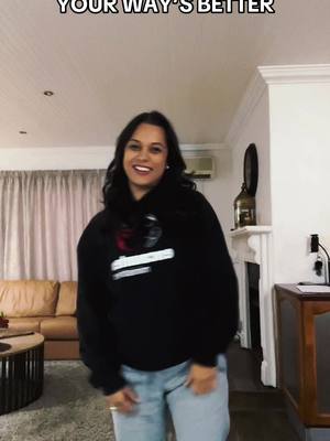 A woman in a black hoodie and blue jeans is smiling and appears to be dancing or posing in a living room. Above her head is text, and behind her is a brown couch, curtains, and a side table.