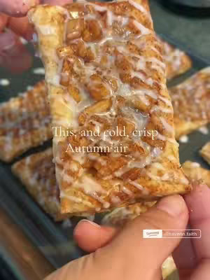 A close-up shot of a rectangular pastry held in a person's hand with additional pastries blurred in the background. The pastry has golden-brown, flaky layers with a topping of a spiced apple and a drizzle of white frosting. The image has a warm, inviting color palette with autumnal colors emphasized.