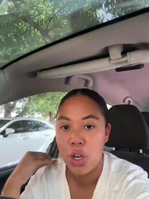 A woman is recording herself in a car, with the camera positioned from a low angle. She's sitting in the driver's seat with the car's interior visible and background includes cars and trees.