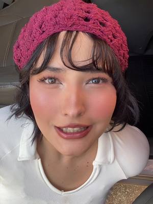 A close-up shot of a young woman's face. She has dark hair with bangs and is wearing a maroon crocheted beret. Her complexion is fair with visible blush on her cheeks. She has dark eyes, and her lips are a deep shade of red. She's wearing a white shirt, and she's looking towards the camera with a subtle smile.