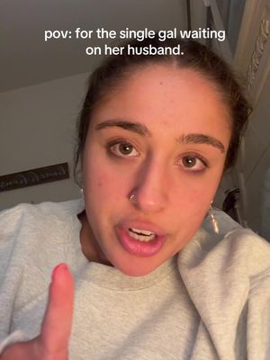 A selfie-style image featuring a woman with brown eyes and her hair pulled back. She's wearing a light gray sweatshirt and a nose ring. She is pointing her right index finger towards the camera and has visible lip gloss. The image has text overlayed at the top that reads "pov: for the single gal waiting on her husband."