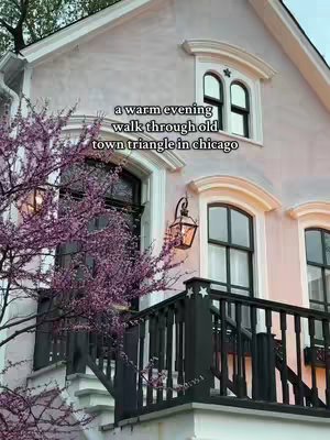 The image captures a Victorian-era pink house with a light-toned exterior, white window frames, and dark windows. Climbing pink flowering trees are on the left. A dark railing and staircase lead up to the front door. A lantern hangs beside the steps.