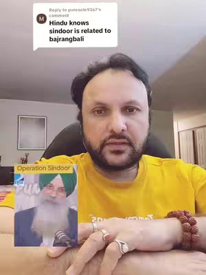 A medium shot features a man with a shaved head, a short beard, and glasses, sitting in front of a neutral background. He is wearing a yellow shirt and has a rudraksha mala on his wrist. In his left hand, he's holding a small, laminated image-in-image depiction. The image is a slightly angled shot. The background is somewhat cluttered. The man is clearly the focus.