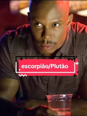 A man with a shaved head is seen seated and looking towards the viewer. He's holding a clear plastic cup with a red liquid. A red and black rectangle overlay with the text "escorpião/Plutão" covers the lower portion of the man's torso. The background is slightly blurred with warm, low lighting.