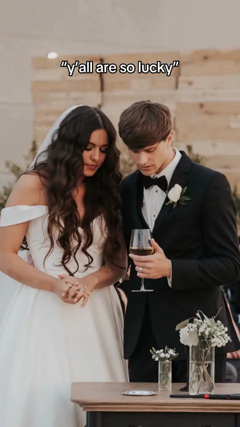 Two people, a woman in a white wedding dress and the man in a black suit, are standing side by side near a wedding table. The woman has a veil on her head and their hands are put together. The man holds a glass with red liquid.