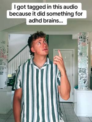 A young man with short, styled hair wearing a striped shirt looks up in the air, pointing upwards with one finger. The background includes what looks like a staircase, a handwritten wall, and a tall cylindrical object covered in text.