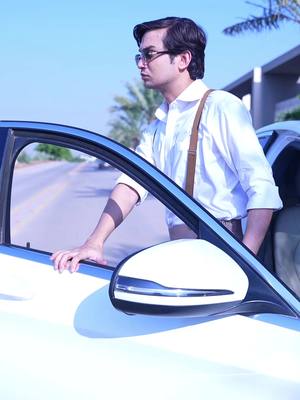 A man in glasses and a white shirt with suspenders leans against a white car, looking to the side. The car door is open, and a road and some greenery are visible in the background.