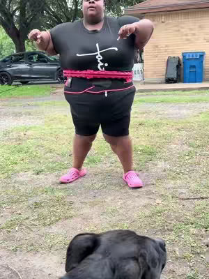 A woman stands outdoors, holding a hula hoop around her waist. She wears a black t-shirt with white text, black shorts, and pink shoes. A black dog is in the front of the frame. The background includes trees, a house, and a parked car.