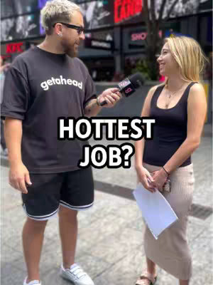 A man holding a microphone is interviewing a woman on a city street. The man is on the left, wearing a black t-shirt and shorts, and the woman is on the right, holding a paper and wearing a black tank top and skirt. A text overlay reads 'Hottest Job?'.