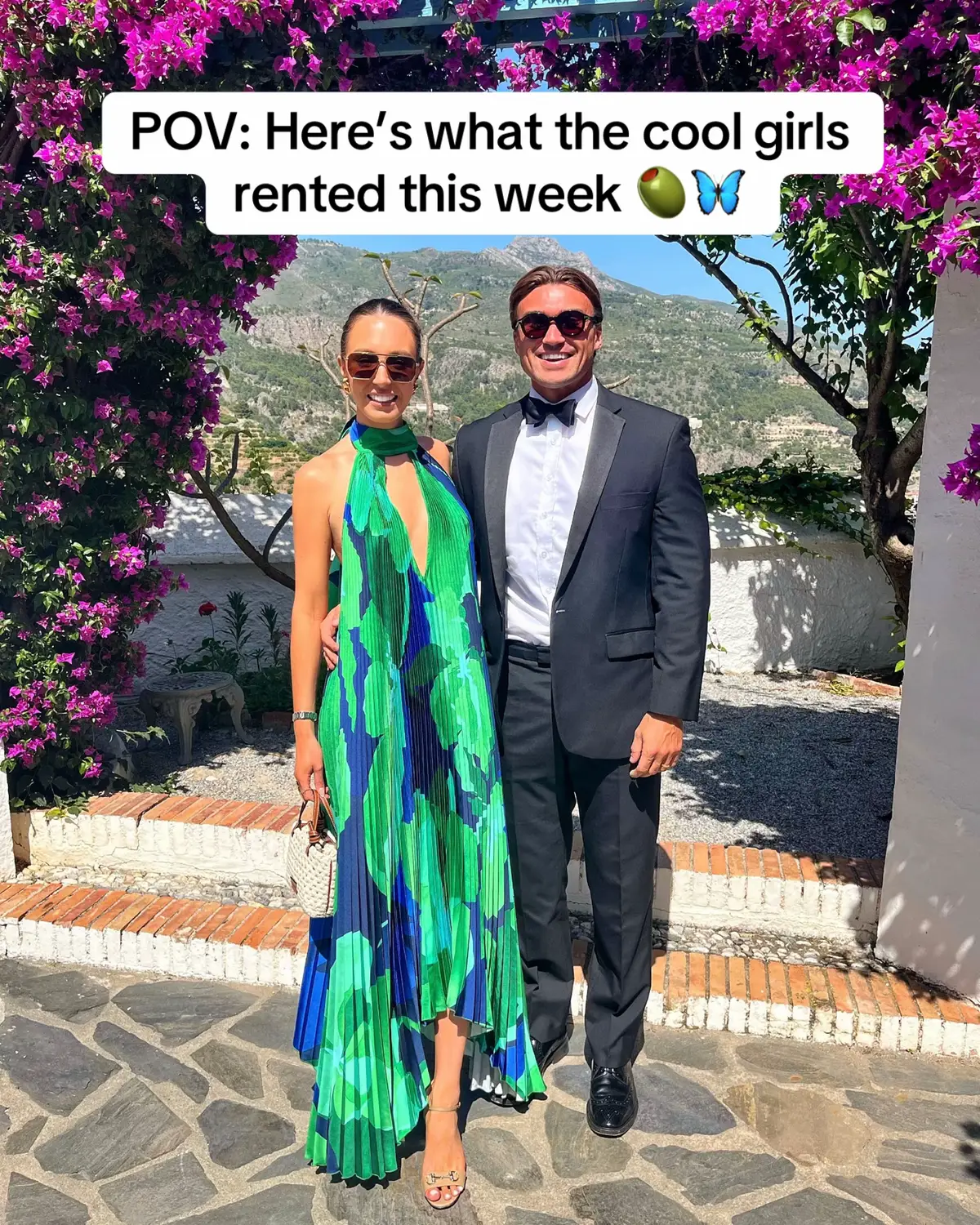 A photo shows a couple posing outside. The woman wears a green and blue patterned dress, sunglasses, and holds a small white purse. The man wears a black suit with a bow tie and sunglasses. They are standing in front of a stone pathway and greenery, with mountains visible in the background. Text overlay reads, 'POV: Here's what the cool girls rented this week'.