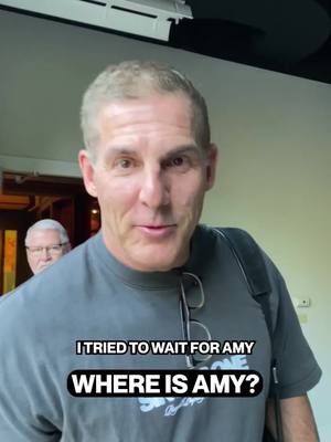A man in a gray t-shirt is centered, looking towards the camera, with the overlay text at the bottom. Behind him, another man with glasses is visible to the left of the frame. The background has neutral tones, suggesting an indoor setting and is slightly out of focus.