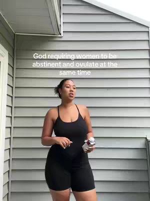 A young Black woman is standing outside, in front of a gray wooden wall. She is wearing a black one-piece outfit with spaghetti straps. She holds a small can in her right hand. The text overlay reads, “God requiring women to be abstinent and ovulate at the same time”.