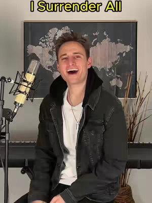 A man sits in front of a microphone, smiling with his mouth open. Behind him is a dark framed picture of a world map with some plants to the right and a keyboard in the background.