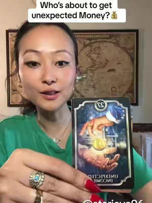 A woman with long dark hair and wearing a green shirt is holding a tarot card in front of her. The card features a gold coin being offered by a hand. Her red nails stand out. She is positioned in front of a blurred background, with a world map on the wall. At the top is a text overlay.