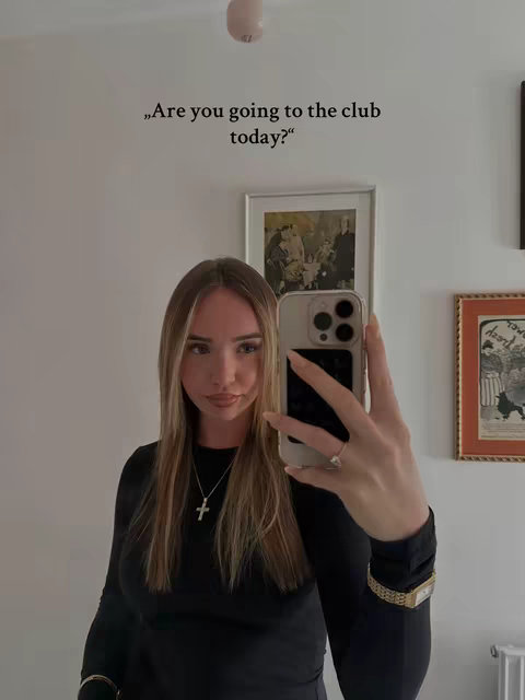 A young woman takes a selfie in front of a white wall. She is holding a phone in her hand and looking at the camera. There is text overlaid on the image stating 'Are you going to the club today?'.