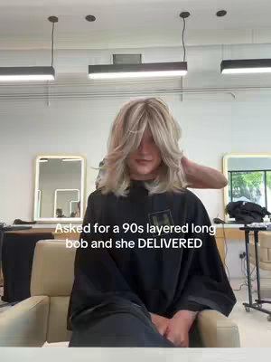 A medium shot shows a person seated in a salon chair with a fresh layered haircut. The person is obscured by their new blonde hair. A stylist's hands are visible, tending to their hair. The background includes salon mirrors, light fixtures, and a bit of the salon's interior.