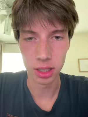 A young man with short brown hair is shown in a close-up shot, facing the camera. He has a neutral expression, with a slight opening of the mouth, as if speaking. The background includes domestic elements. The lighting is consistent with natural light, the overall composition is simple and direct.