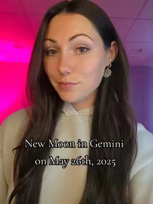 A woman with long brown hair is centered. She wears a cream-colored hoodie, and a small diamond-like earring. Text overlay states, 'New Moon in Gemini on May 26th, 2025'. The background gradient is mainly pink and purple.