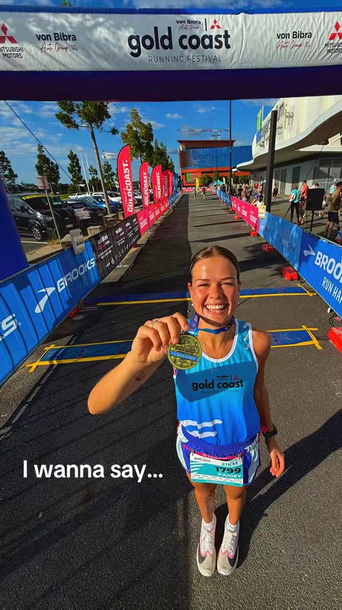 A woman in a blue athletic top and shorts smiles while holding up a medal at a running event. The background shows a finish line beneath an arch and crowd of people