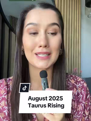 A woman with long, dark hair, wearing a patterned top, looks directly at the camera and speaks. The background includes a blurred interior with wooden paneling. A white rectangular overlay at the bottom of the frame features the text 'August 2025 Taurus Rising' in a black sans-serif font. A small microphone is positioned in front of her mouth.