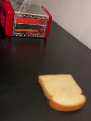 A slice of bread in the foreground, positioned to the right. In the background is a red toaster on a black table, and white vapor visible indicating the toasting process. The background is an off-white wall.