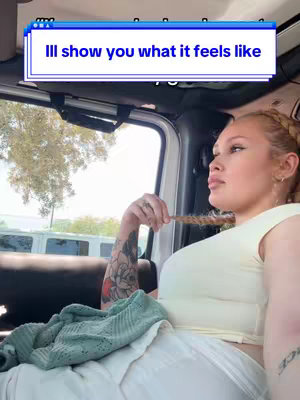 A woman with blonde hair, wearing a light yellow top and white pants, sitting inside a car. She's turned towards the right, looking off-camera, with a thoughtful expression. A hand with a tattoo holds a strand of her hair. The background is a blurred view of outside through the car window.