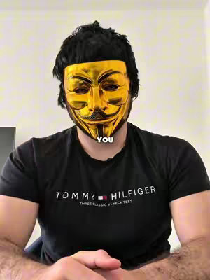 A man wearing a golden Guy Fawkes mask gives a direct gaze. He's wearing a black t-shirt which has the Tommy Hilfiger logo. At the bottom of the mask and in white text is the word "YOU". The background is a plain, neutral wall.