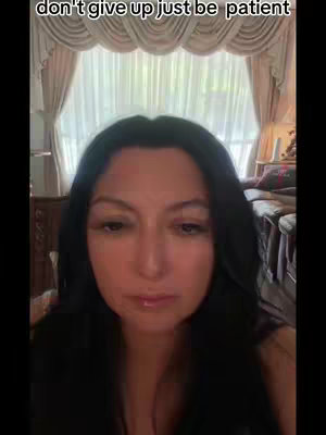 A woman with dark hair and a neutral expression is centered in the frame. The background includes a glimpse of a window with curtains and a cozy interior setting.