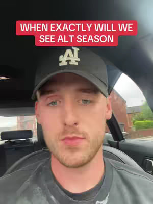 A selfie-style shot with a man sitting in a car. The man is fair-skinned with a short beard and short blonde hair. He is wearing a black baseball cap with a white 'A' emblem, a dark shirt, and has a serious expression. The background is blurred, showing car interior and a building.
