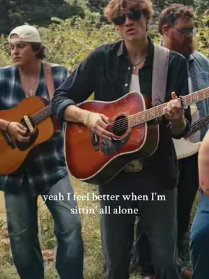 A group of young men playing guitars outdoors, likely in a park or natural setting. The central figure is playing an acoustic guitar and wearing sunglasses. Text overlays the scene.