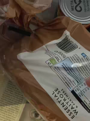 The image shows a close-up of a package, likely food, lying on top of crumpled plastic wrapping. Visible text includes the brand name "ESSENTIAL WAITROSE & PARTNERS" and nutritional information with some of the packaging details. The background is blurred and appears to contain a can and possibly other items within a cupboard or storage space.