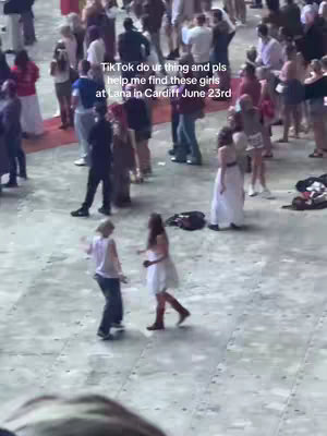 A candid shot taken from a slightly elevated perspective, showing a large outdoor space filled with a crowd of people. Two young women are in the foreground, engaged in a conversation or interaction. The backdrop is blurred with people, and the scene has a natural, unedited feel, possibly at a concert or event.