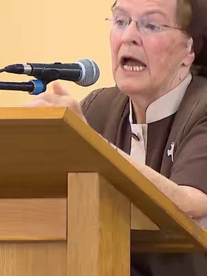An image featuring Sr. Briege McKenna speaking from behind a wooden podium. She is wearing glasses and a brown habit over a white collar. A microphone is positioned near her mouth.