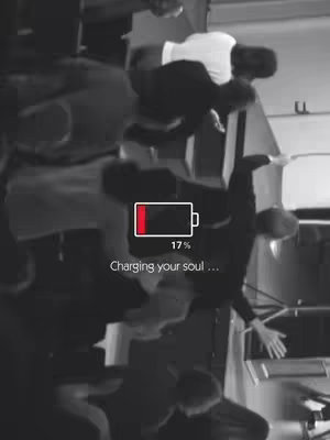 A grayscale image shows a crowd of people, possibly at an event or performance. A large battery icon is positioned at the lower center, with a red bar indicating 17% charge. The text 'Charging your soul...' appears below the battery.