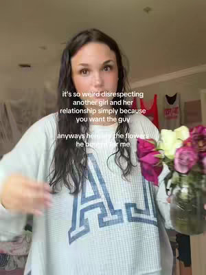 A woman stands indoors, holding a vase of flowers. The background is blurred, with hints of a room and some clothing articles on display. The woman is wearing a light-colored top with a large letter 'A' on it, and the text overlayed onto the image conveys a message about relationships.