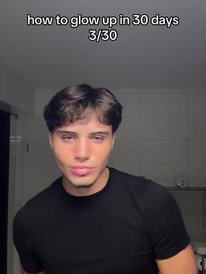 A young man with brown hair and a black t-shirt is centered in the frame. Text overlays appear above: 'how to glow up in 30 days 3/30'. The background is a dimly lit bathroom with a light fixture and a glimpse of a sink.