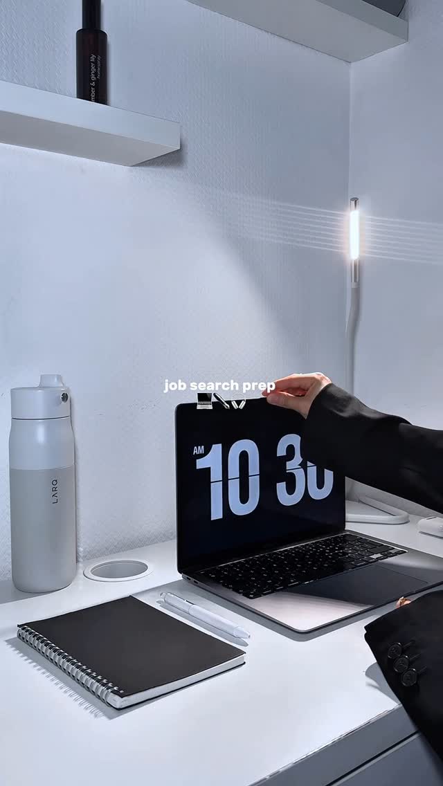 A minimalist desk setup with a white surface. A laptop displaying a clock, a notebook, pen, and water bottle are visible. A hand is placed on the laptop, and a jacket is on the desk. The image is brightly lit with focused lighting on objects.