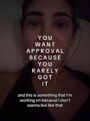 A selfie-style video cover image. The creator, a woman, is centered in the frame, looking directly at the camera. Text overlayed on her face reads: "YOU WANT APPROVAL BECAUSE YOU RARELY GOT IT". She's in a dimly lit setting wearing a dark top, with an intimate or casual vibe. The background appears soft, indicating it might be indoors, likely a bedroom setting.