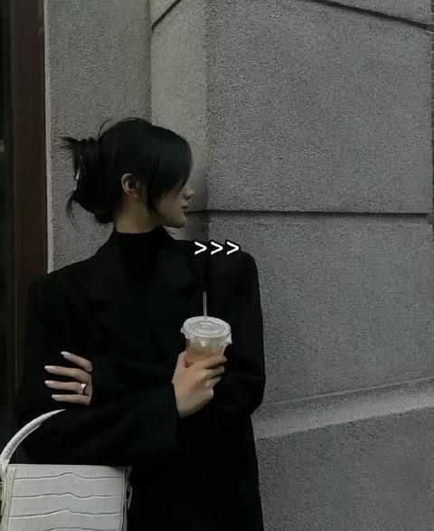 A woman in a black blazer and turtleneck, holding a disposable coffee cup, leans against a gray concrete wall. She looks away from the camera, with her hair pulled back.