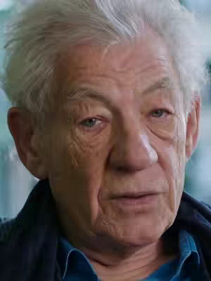 Close-up shot of an elderly man with white hair, looking directly at the camera. His facial features are prominent, with wrinkles and lines emphasizing his age. The background is blurred, suggesting an indoor setting.