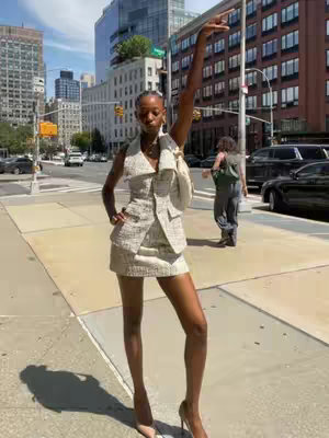 A woman stands on a city sidewalk. She is wearing a light-colored suit and high heels and has one arm raised up with her hand open. The background features city buildings, trees, and other people walking.